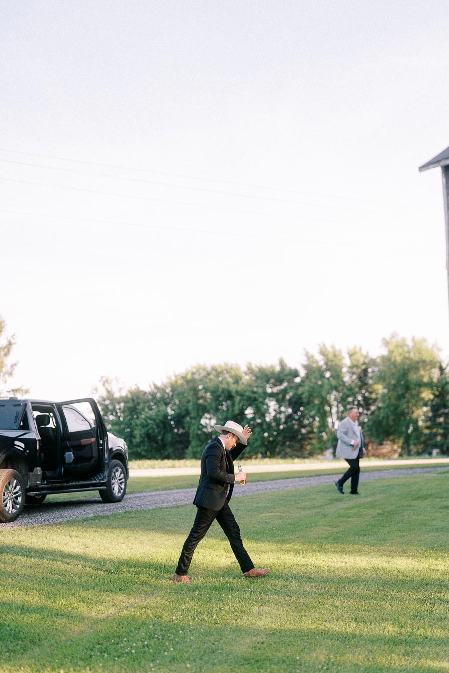 Man in a cowboy hat walking across a lawn near a black pickup truck during an outdoor farm wedding in Chatham-Kent, Ontario.