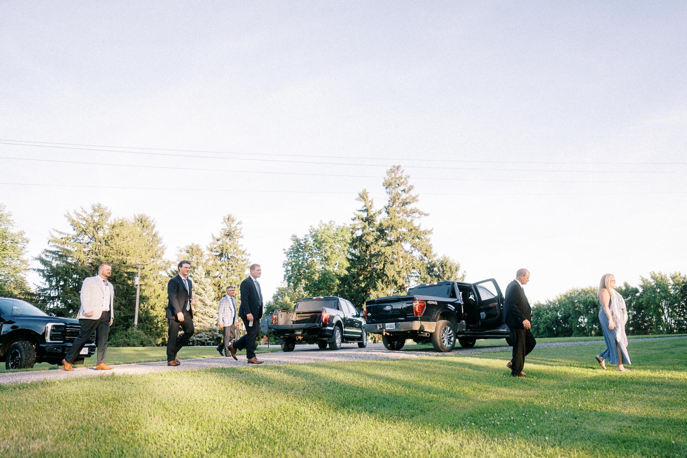 Wedding guests arrive on a private farm estate lawn near Chatham-Kent, Ontario, with black pickup trucks parked along a gravel driveway.