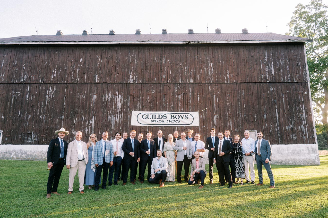 Wedding party group portrait on a grassy farm lawn in front of a rustic wooden barn near Chatham-Kent, Ontario, Canada.
