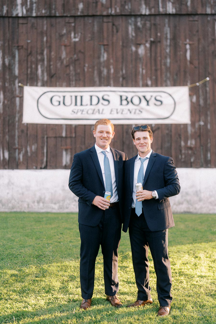 Two men in dark suits holding cans on grass in front of a rustic barn with a Guilds Boys Special Events banner near Chatham-Kent, Ontario.