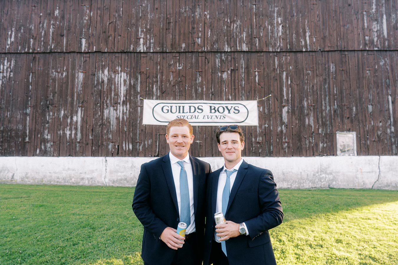 Two men in suits holding cans on a lawn in front of a rustic barn wall with a Guilds Boys Special Events sign near Chatham-Kent, Ontario.