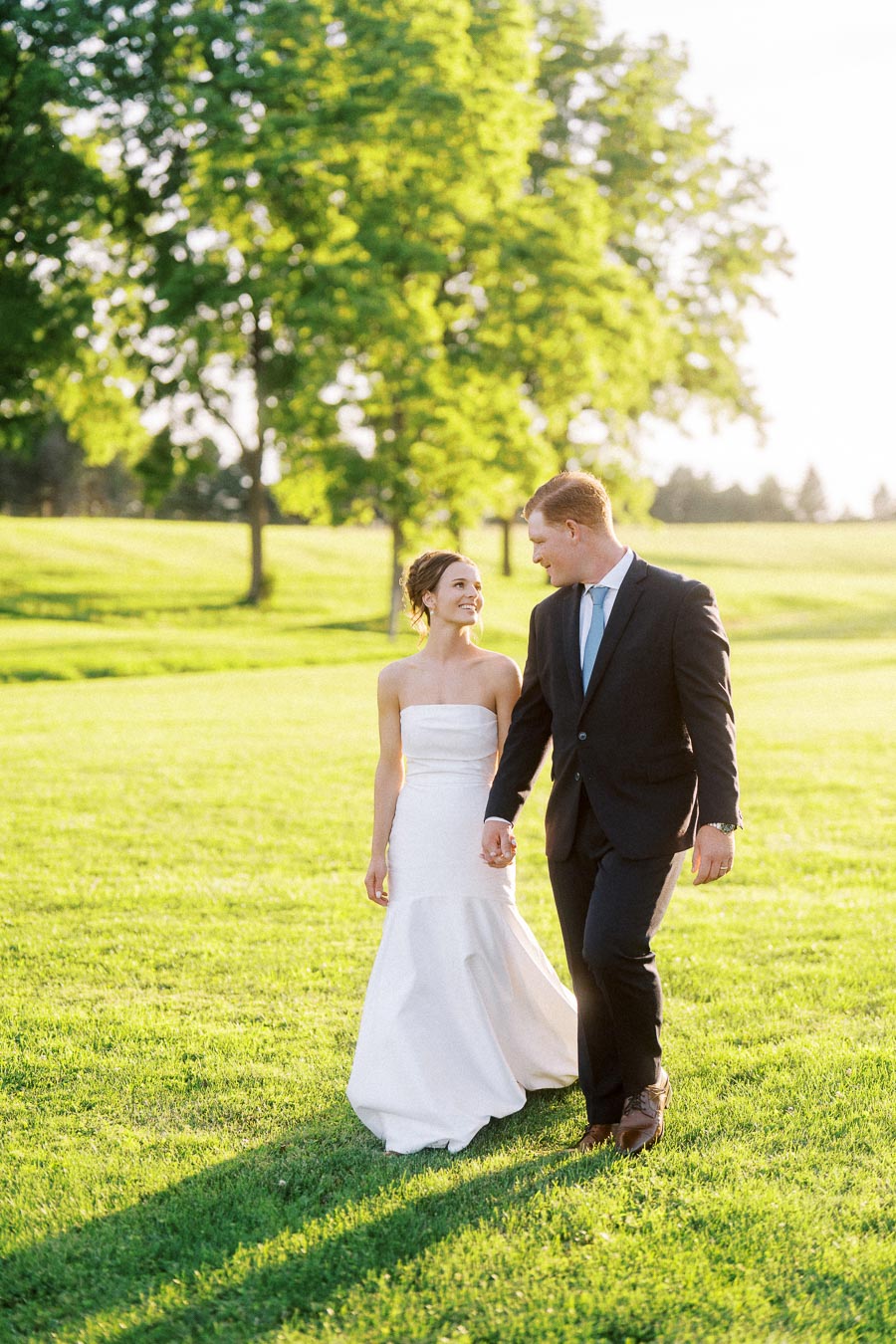 Bride and groom walking hand in hand on a sunny green lawn during an outdoor farm wedding near Chatham-Kent, Ontario, Canada.