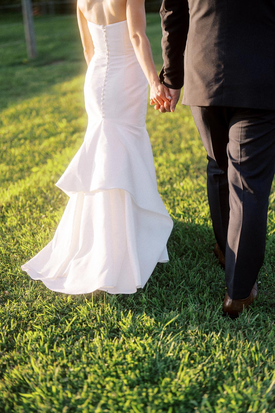 Bride in a white gown and groom in a black suit holding hands while walking across sunlit grass at an outdoor farm wedding near Chatham-Kent, Ontario.