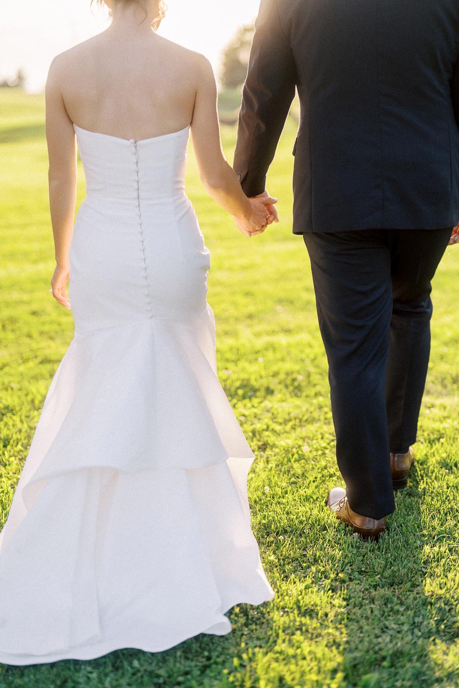 Bride in a white strapless gown and groom in a dark suit walk hand in hand across a sunlit grassy field near Chatham-Kent, Ontario.