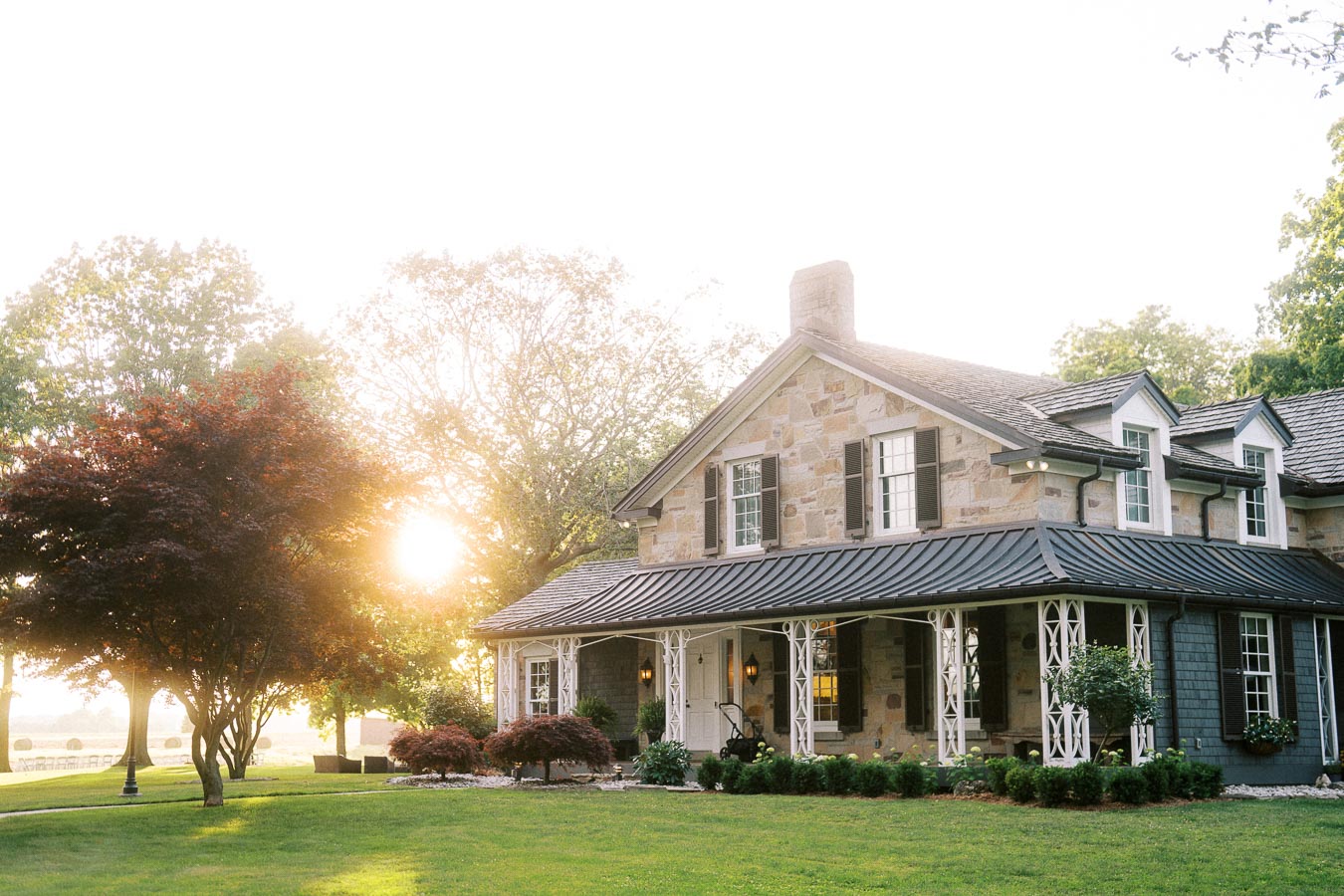 Stone farmhouse on a private estate lawn at sunset near Chatham-Kent, Ontario, Canada, with trees, porch, garden shrubs, and warm outdoor light.