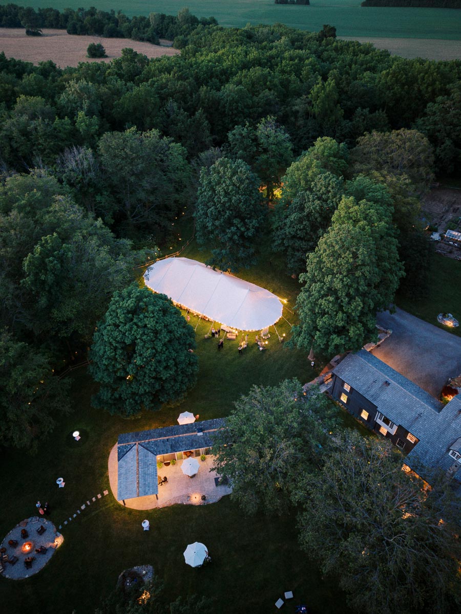Aerial view of a white wedding tent glowing at dusk on a private farm estate near Chatham-Kent, Ontario, surrounded by trees, fields, lawn, and house.