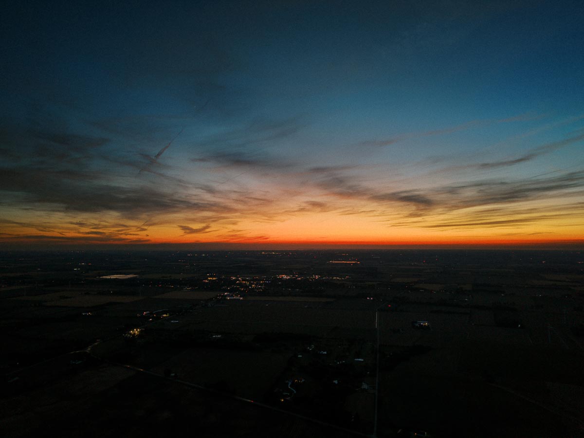 Aerial sunset view over rural farmland near Chatham-Kent, Ontario, with dark fields, distant lights, and colorful evening clouds.