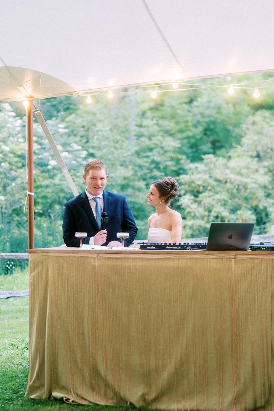 Bride and groom stand behind a draped DJ table under a wedding tent with string lights during an outdoor farm wedding near Chatham-Kent, Ontario.