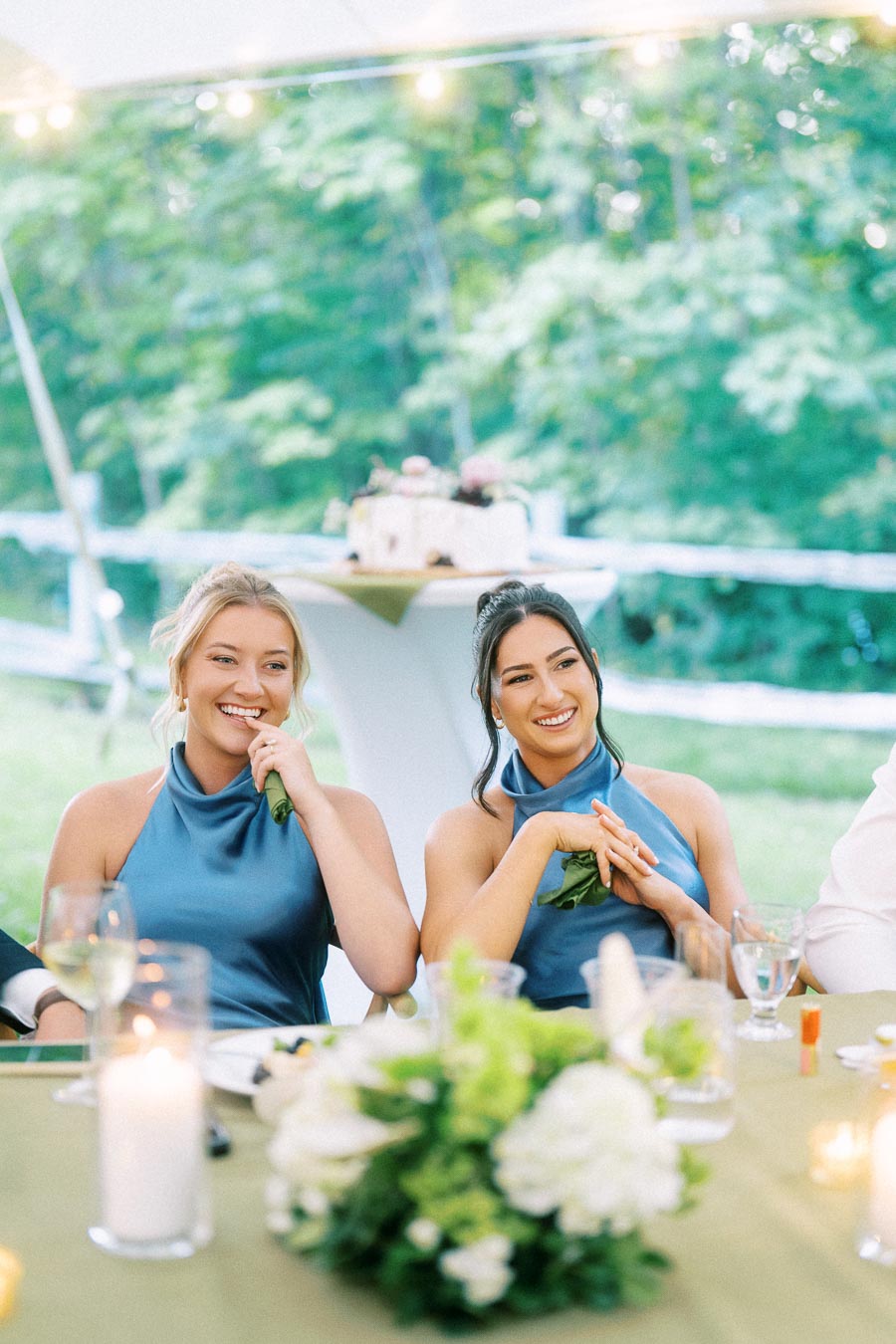 Two bridesmaids in blue dresses smiling at a tented outdoor wedding reception table with candles, flowers, and glasses in Chatham-Kent, Ontario.