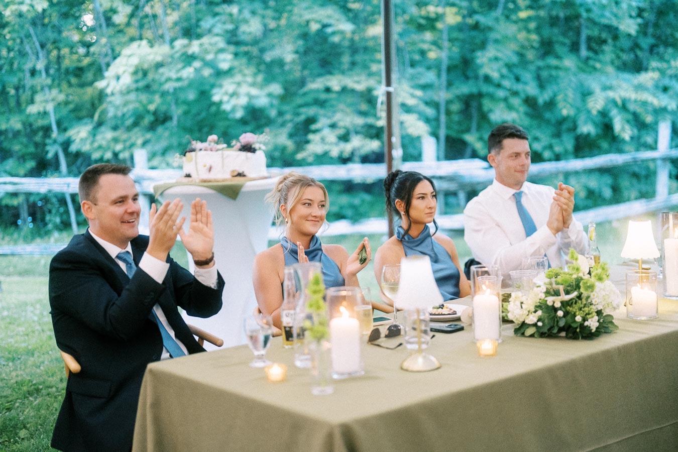 Wedding guests applauding during an outdoor private estate reception dinner near Chatham-Kent, Ontario, with candles, flowers, and woodland greenery.