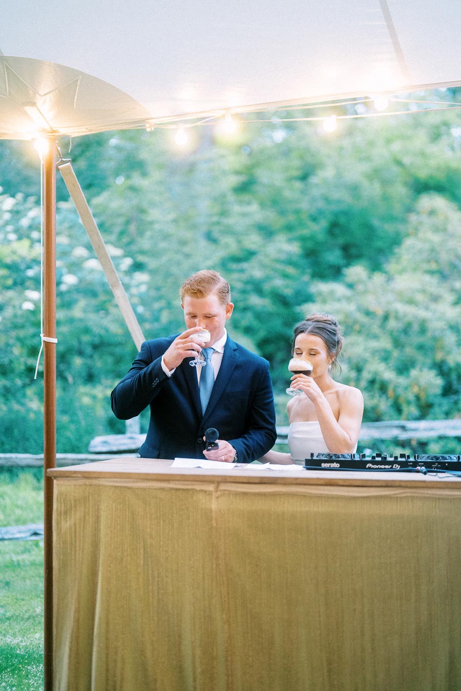 Bride and groom drink from glasses under a lit wedding tent at a private outdoor farm reception near Chatham-Kent, Ontario, Canada.