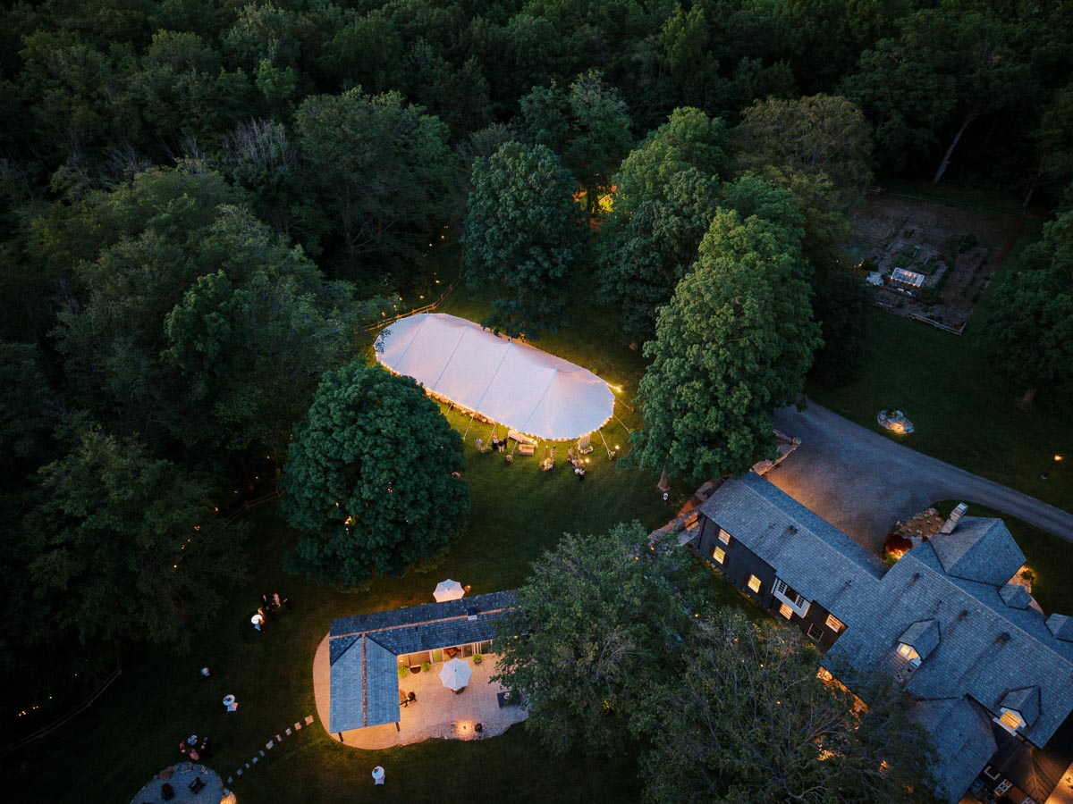 Aerial dusk view of a glowing white wedding tent on a wooded private estate near Chatham-Kent, Ontario, with farmhouse, patio, lawn, trees, and guests.