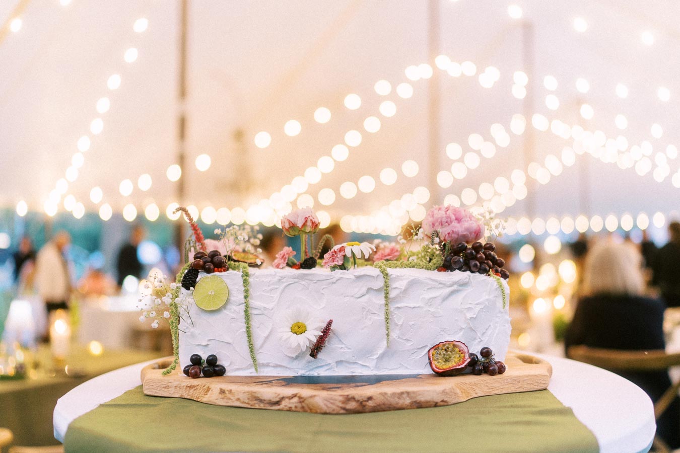 Decorated wedding cake with flowers and fruit on a wood board inside a glowing tented farm reception near Chatham-Kent, Ontario.