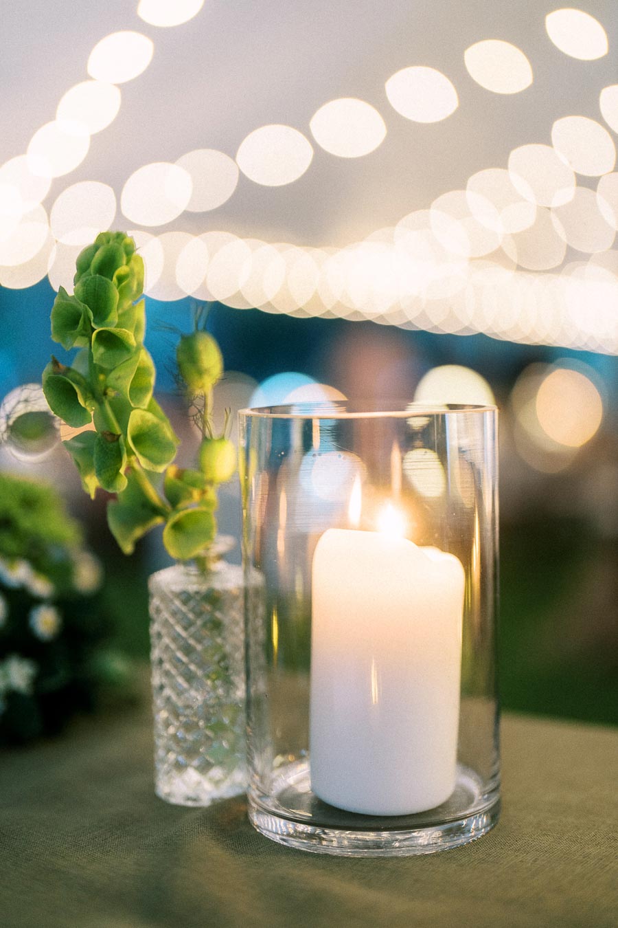 Lit white candle in a glass hurricane vase with green floral decor and warm string lights at an outdoor tented wedding near Chatham-Kent, Ontario.