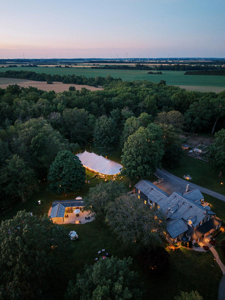 Aerial view of a tented outdoor wedding dinner on a private farm estate near Chatham-Kent, Ontario, surrounded by trees and fields at dusk.