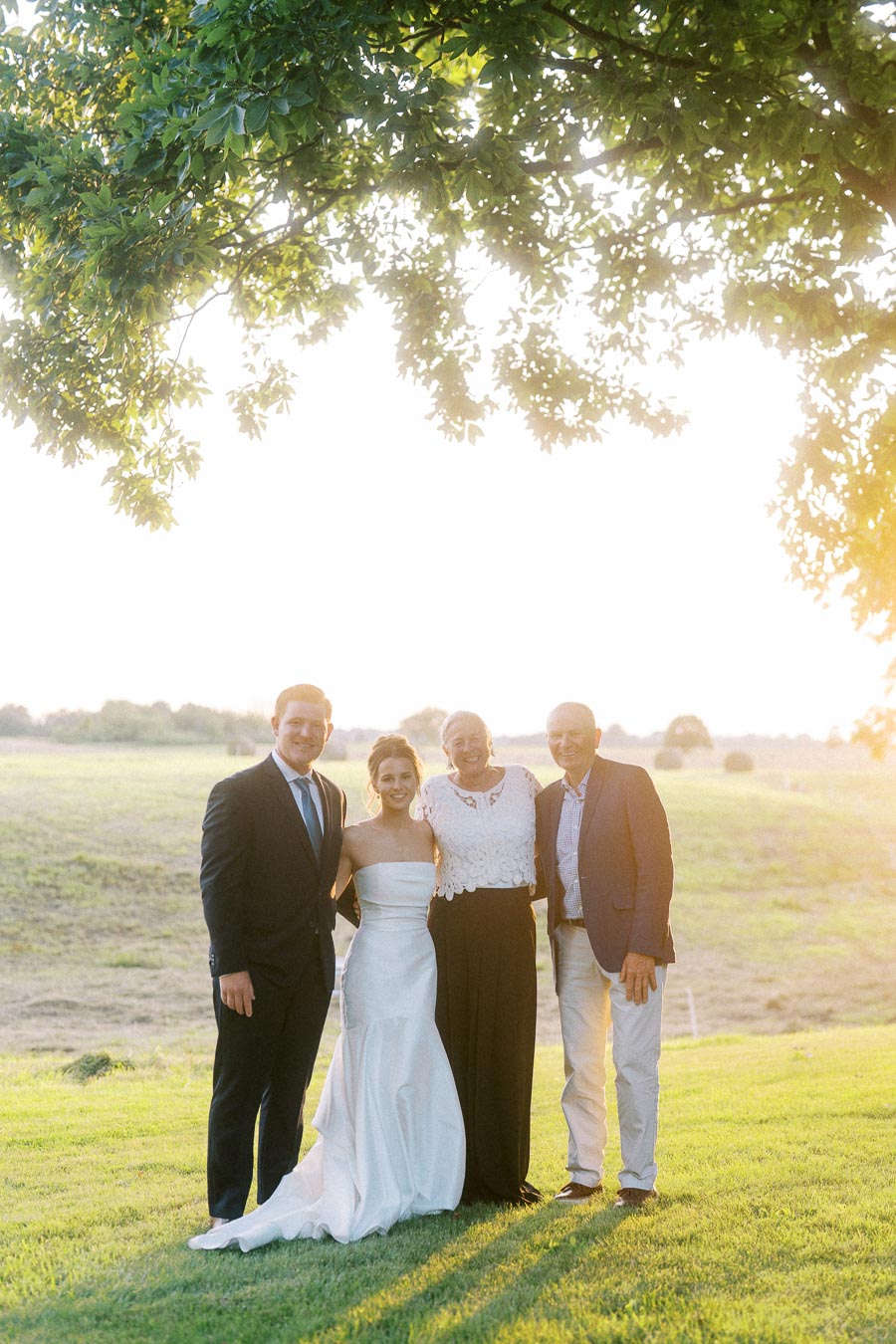 Bride, groom, and two family members posing outdoors on a farm lawn near Chatham-Kent, Ontario, at golden hour under tree branches.
