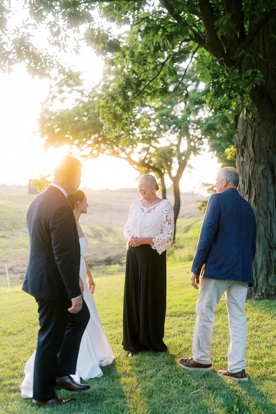 Bride, groom, and guests talking under a large tree during a golden hour outdoor farm wedding near Chatham-Kent, Ontario, Canada.