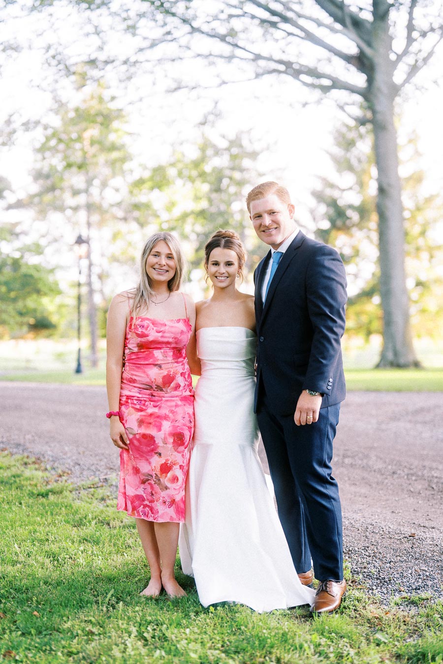 Bride posing with two wedding guests on a grassy farm property near Chatham-Kent, Ontario, with trees and a gravel drive behind them.