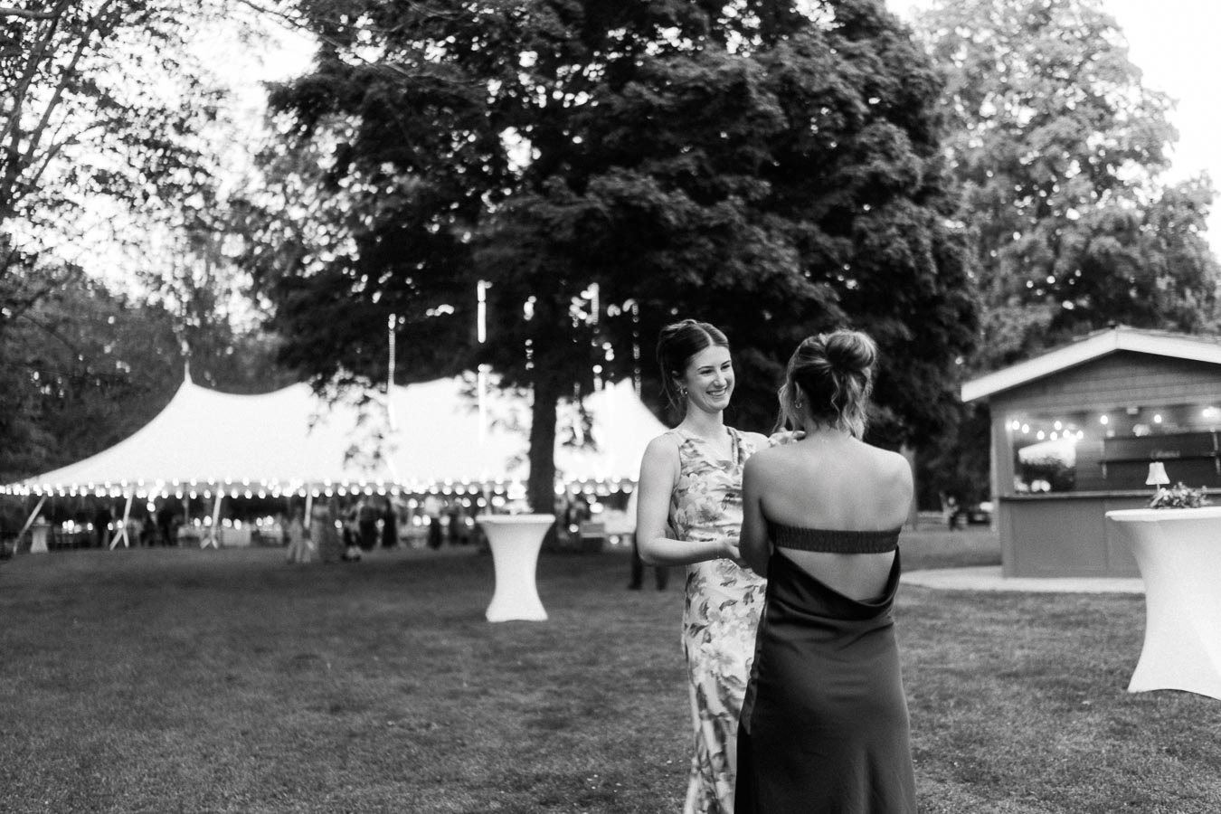 Black and white outdoor wedding reception on a private estate near Chatham-Kent, Ontario, with two women talking beside a lit tent on a farm lawn.