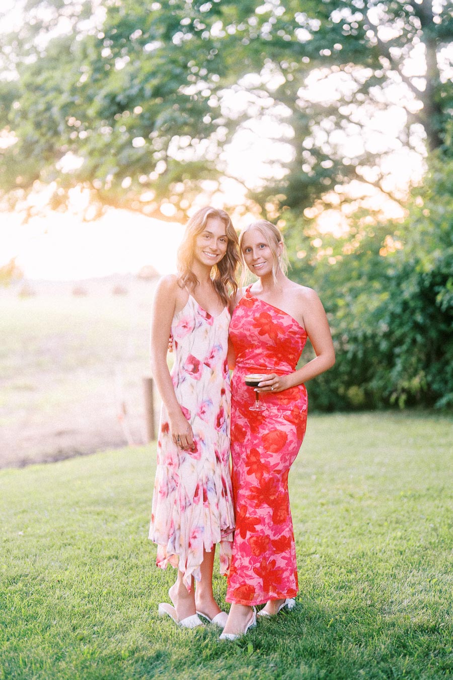 Two women in floral dresses posing on a grassy lawn at an outdoor wedding near Chatham-Kent, Ontario, with trees and sunset light.