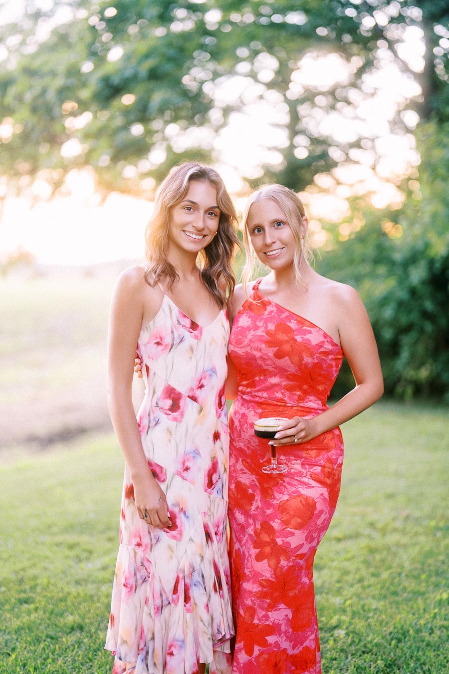 Two women in floral dresses posing outdoors on a grassy private estate near Chatham-Kent, Ontario during a summer wedding celebration.