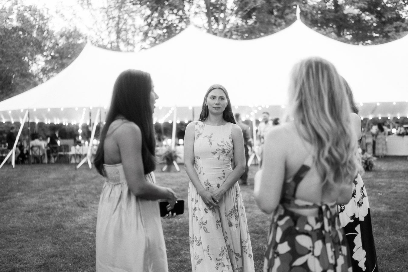 Women talking on grass outside a lit wedding tent during an outdoor private estate farm wedding near Chatham-Kent, Ontario, Canada.