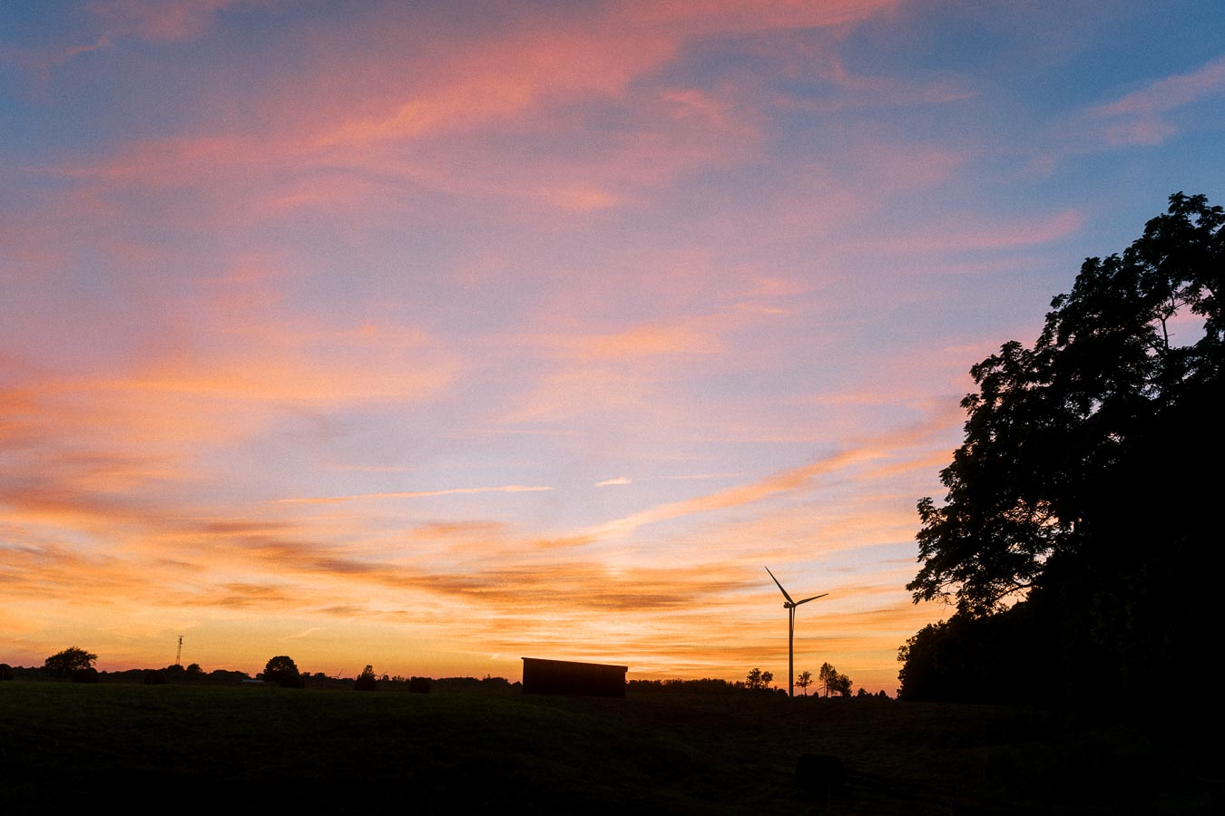 Colorful sunset sky over a rural private farm field near Chatham-Kent, Ontario, Canada, with silhouetted trees, barn structure, and wind turbine.