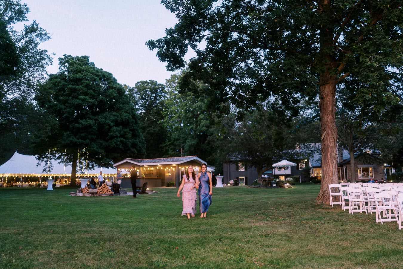 Outdoor farm wedding reception on a private estate near Chatham-Kent, Ontario, with a lit tent, lawn seating, trees, and guests walking at dusk.