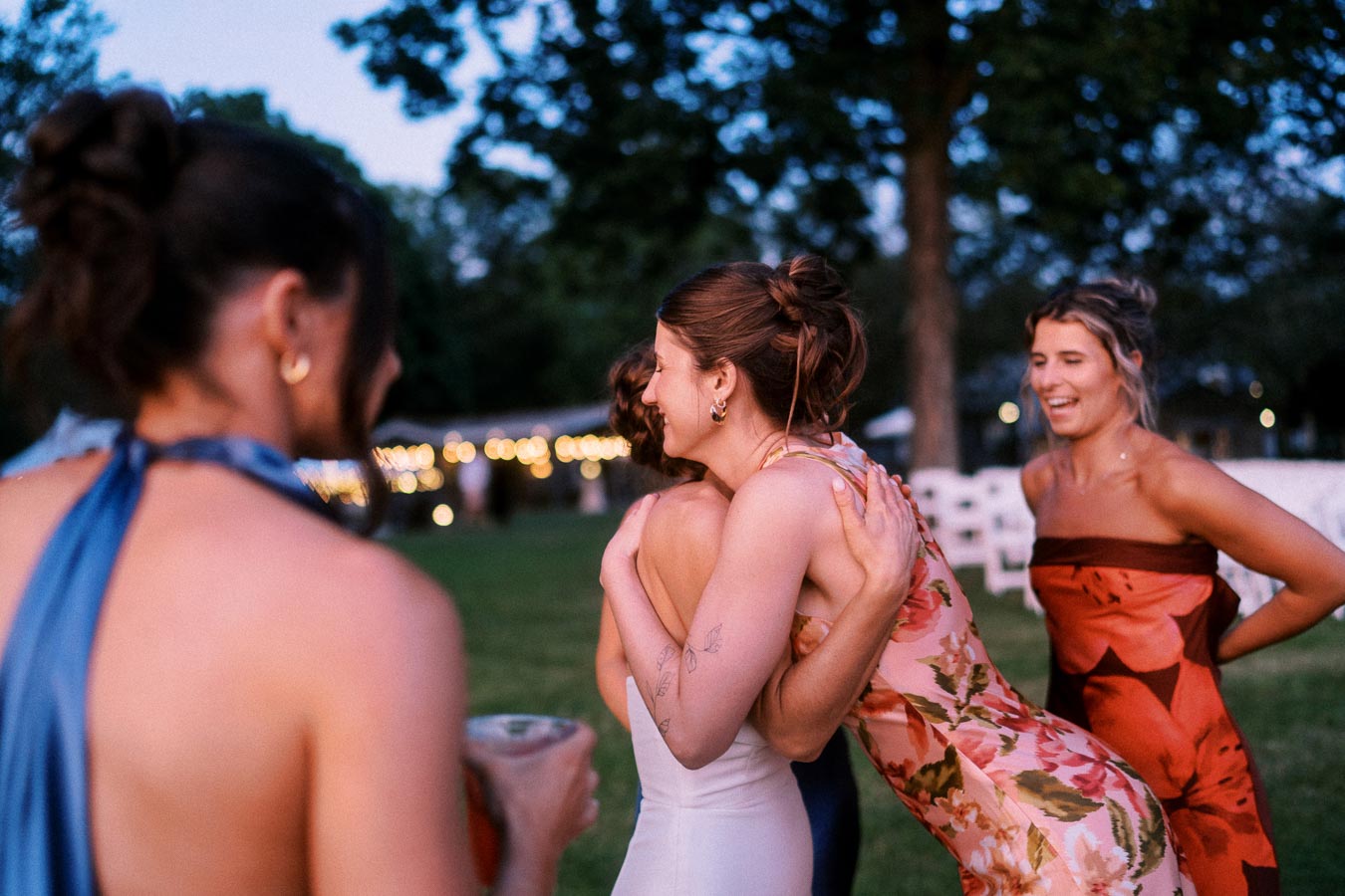 Wedding guests hug and talk on a grassy private estate near Chatham-Kent, Ontario, with a tent, string lights, white chairs, and trees at dusk.