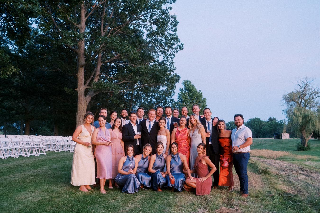 Wedding guests pose for a group portrait outdoors on a farm estate near Chatham-Kent, Ontario, with white ceremony chairs and trees.