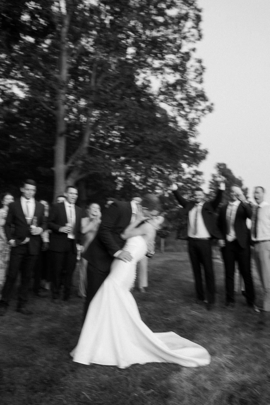 Black and white outdoor wedding photo of a groom dipping and kissing the bride in a white gown while guests cheer on a grassy private estate near Chatham-Kent, Ontario.