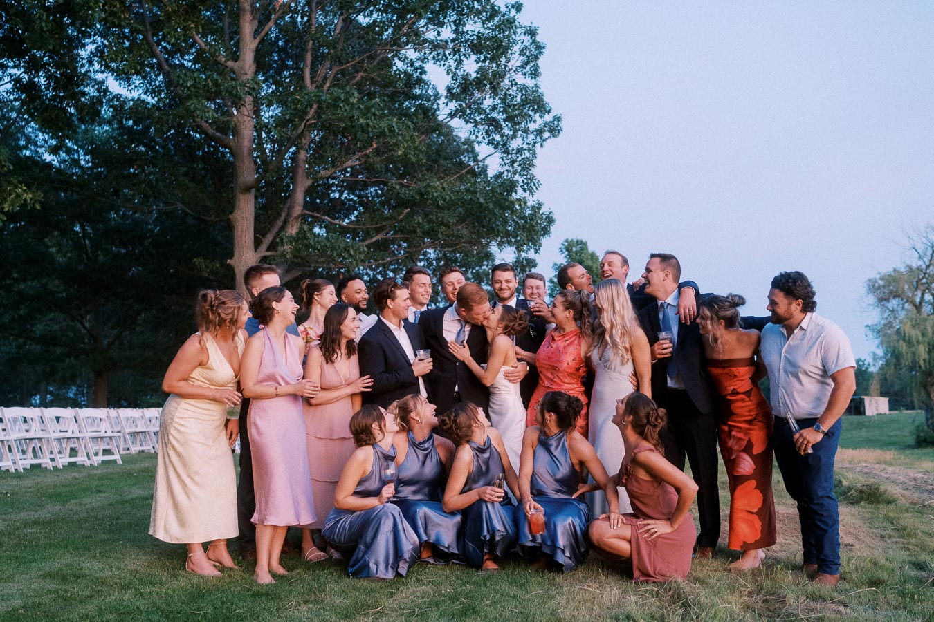 Wedding party laughing together outdoors on a farm estate lawn near Chatham-Kent, Ontario, with trees, grass, and white ceremony chairs.