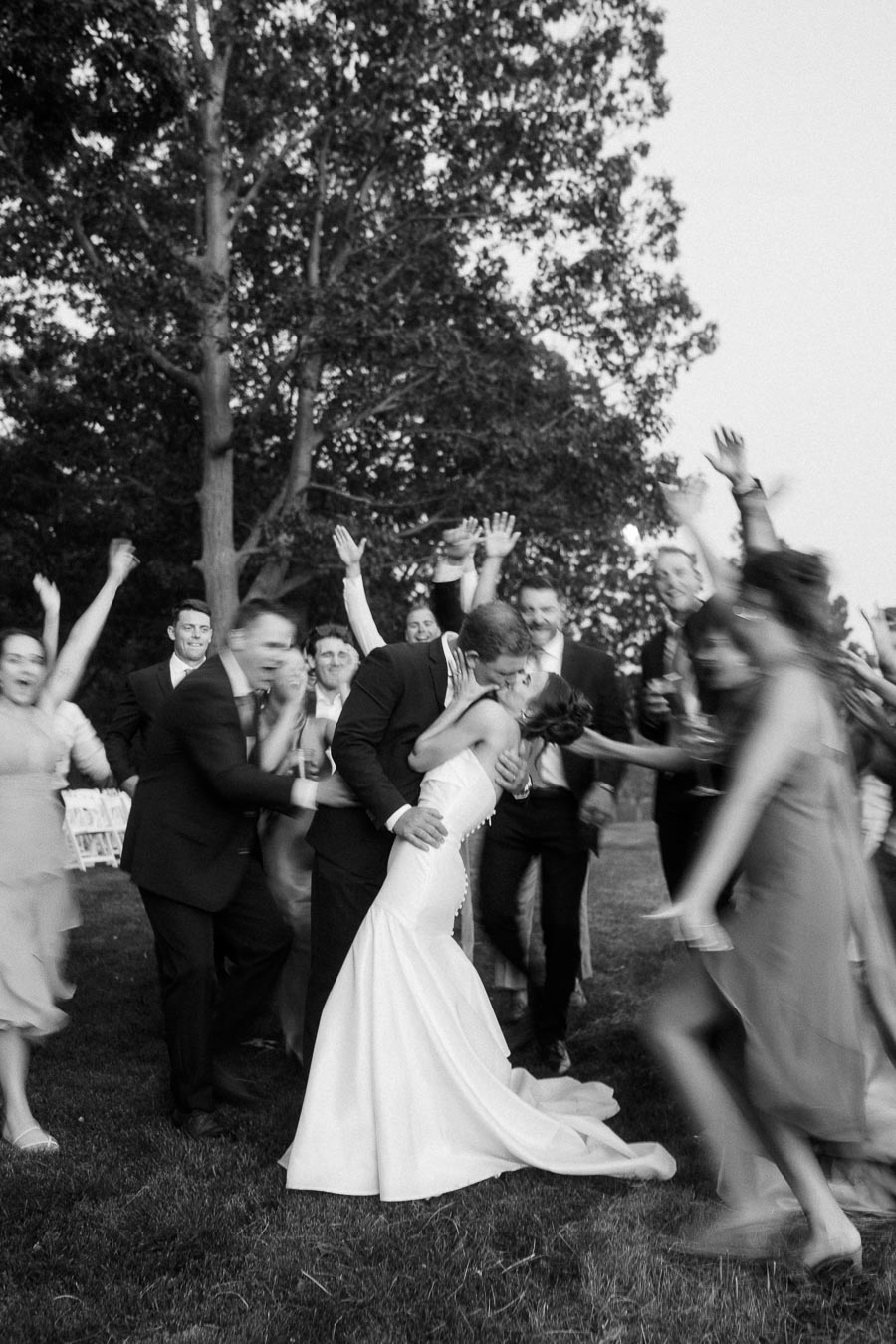 Black and white outdoor wedding photo of bride and groom kissing while cheering guests celebrate on a farm lawn near Chatham-Kent, Ontario.