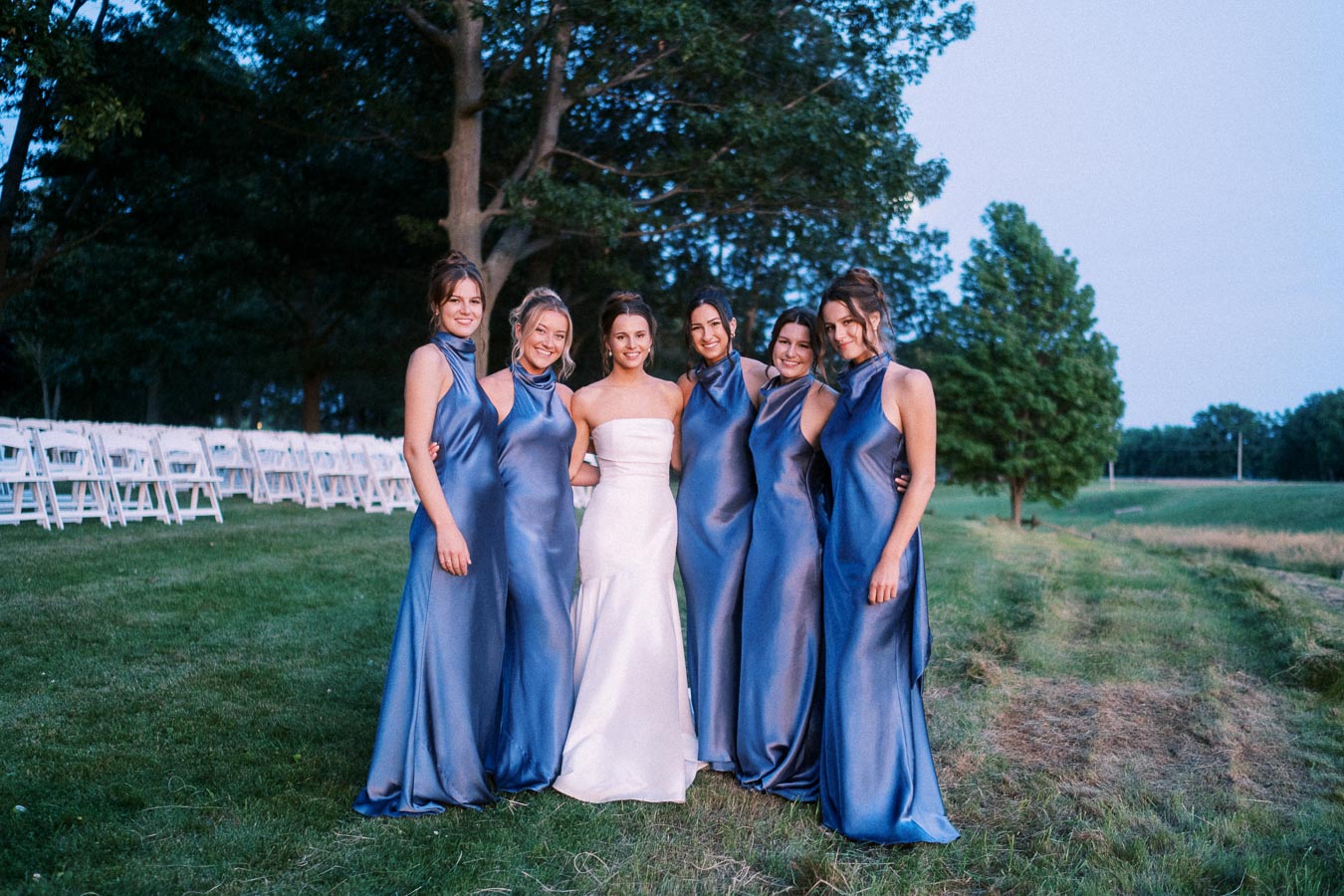 Bride with bridesmaids in blue dresses posing outdoors beside white wedding chairs on a private farm estate near Chatham-Kent, Ontario.