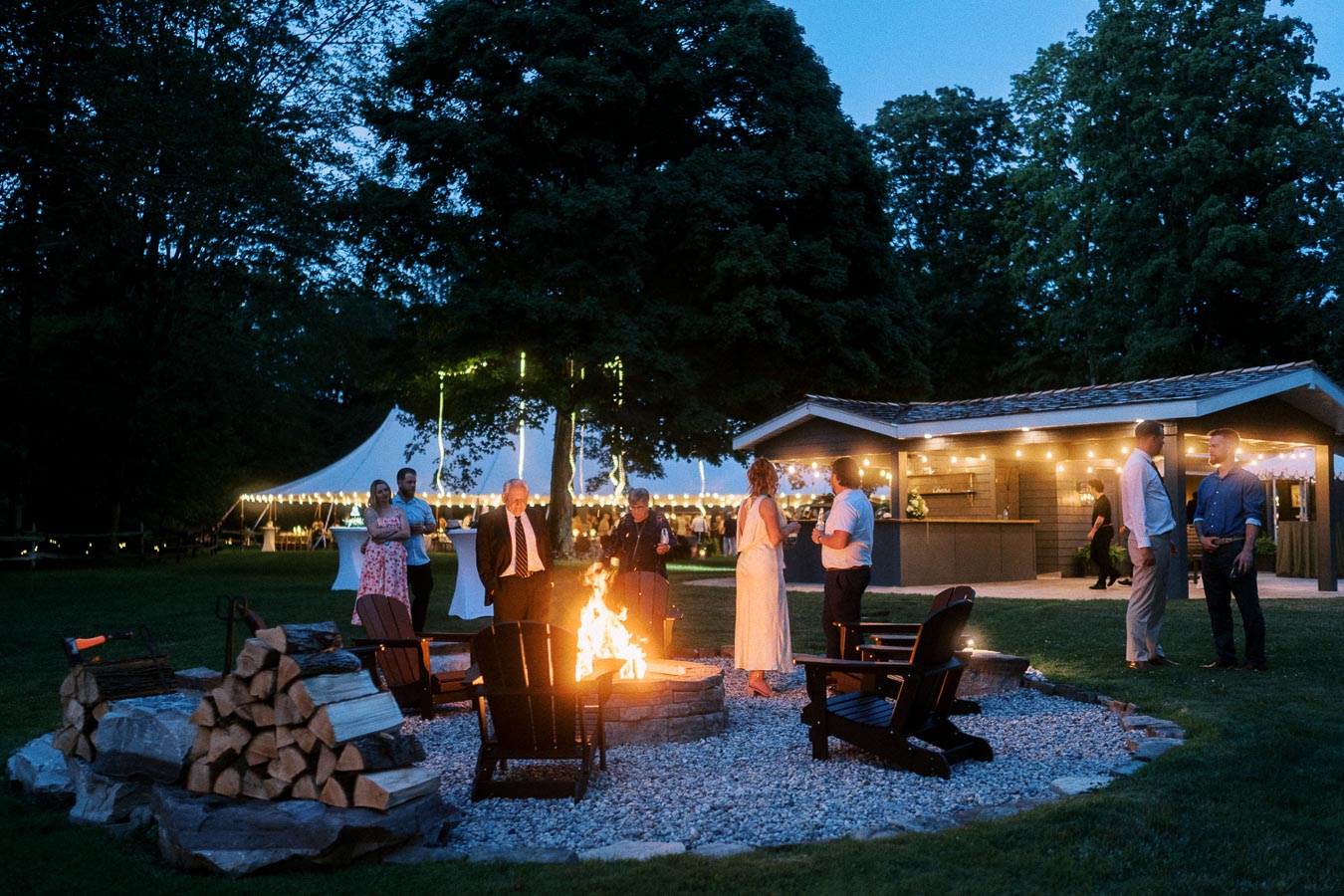 Outdoor private estate wedding reception near Chatham-Kent, Ontario, with guests gathered around a fire pit beside a tent and illuminated pavilion at dusk.