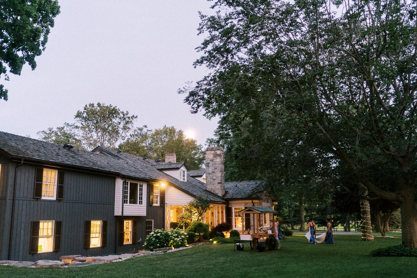 Outdoor wedding guests on a lawn beside a lit private estate house near Chatham-Kent, Ontario, Canada, with large trees at dusk.