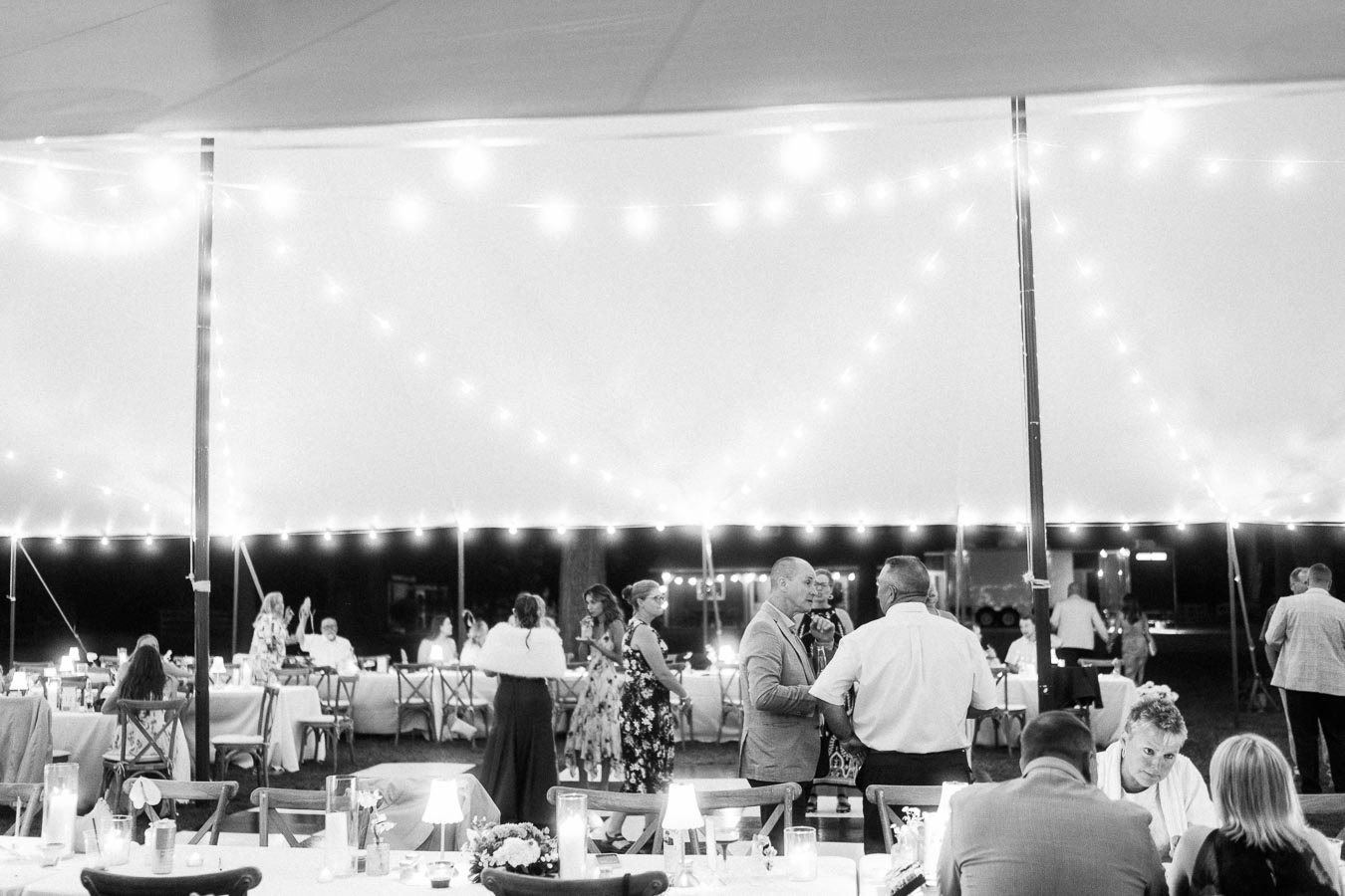 Black and white photo of guests dining and talking under a lit wedding tent at a private estate farm wedding near Chatham-Kent, Ontario.