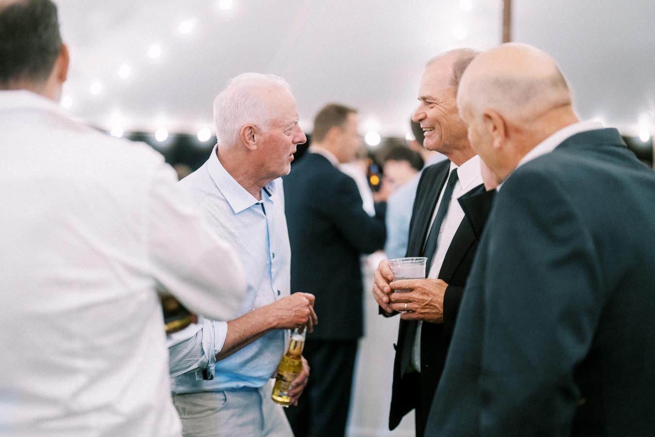 Male wedding guests talking and holding drinks inside a lit reception tent near Chatham-Kent, Ontario, Canada.
