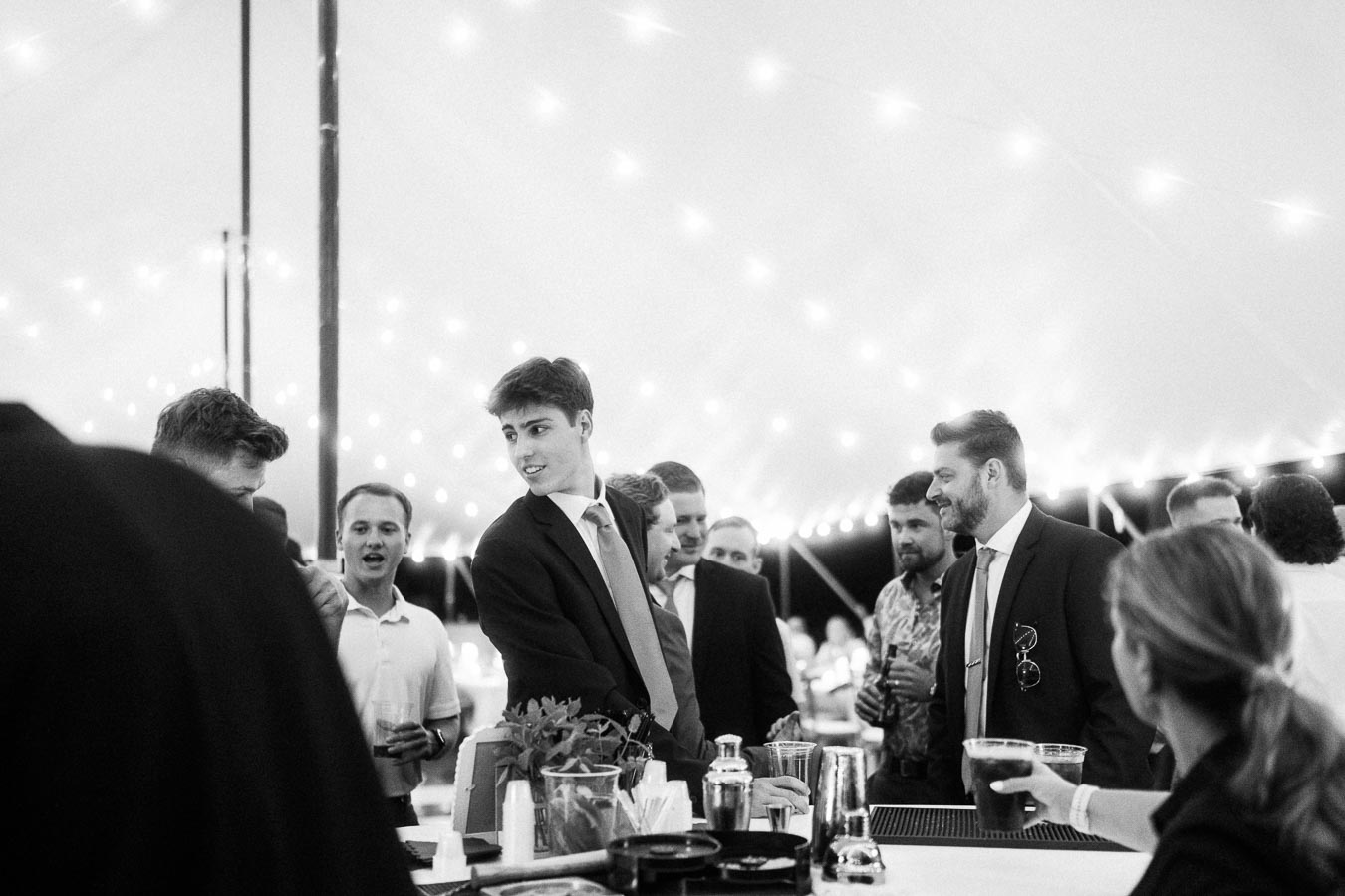Black and white photo of wedding guests gathered at a bar inside a lighted reception tent near Chatham-Kent, Ontario.