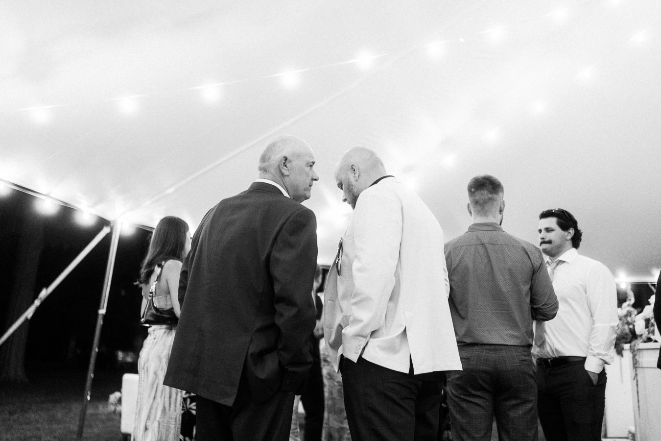 Black and white photo of wedding guests talking under a lit reception tent at a private estate near Chatham-Kent, Ontario.