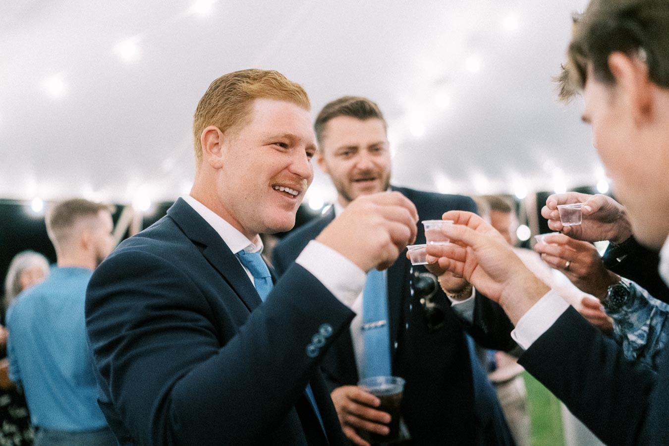 Wedding guests in suits raising small cups for a toast inside a lit reception tent at a private farm estate near Chatham-Kent, Ontario.