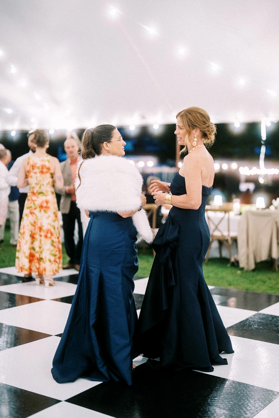 Two women in navy formal dresses talk on a black and white dance floor under a lit wedding tent near Chatham-Kent, Ontario.