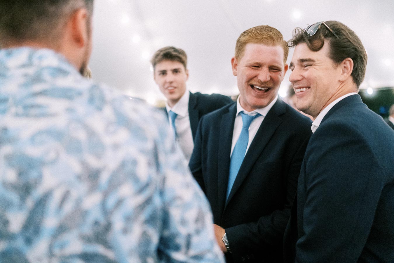 Men in suits laughing and talking under a wedding tent with string lights near Chatham-Kent, Ontario, Canada.