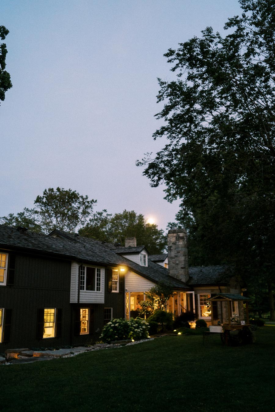 Evening view of a private estate farmhouse with lit windows, stone chimney, lawn, garden shrubs, and trees near Chatham-Kent, Ontario.