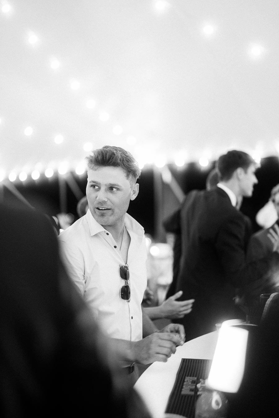 Black and white candid photo of wedding guests talking under a reception tent with string lights near Chatham-Kent, Ontario, Canada.