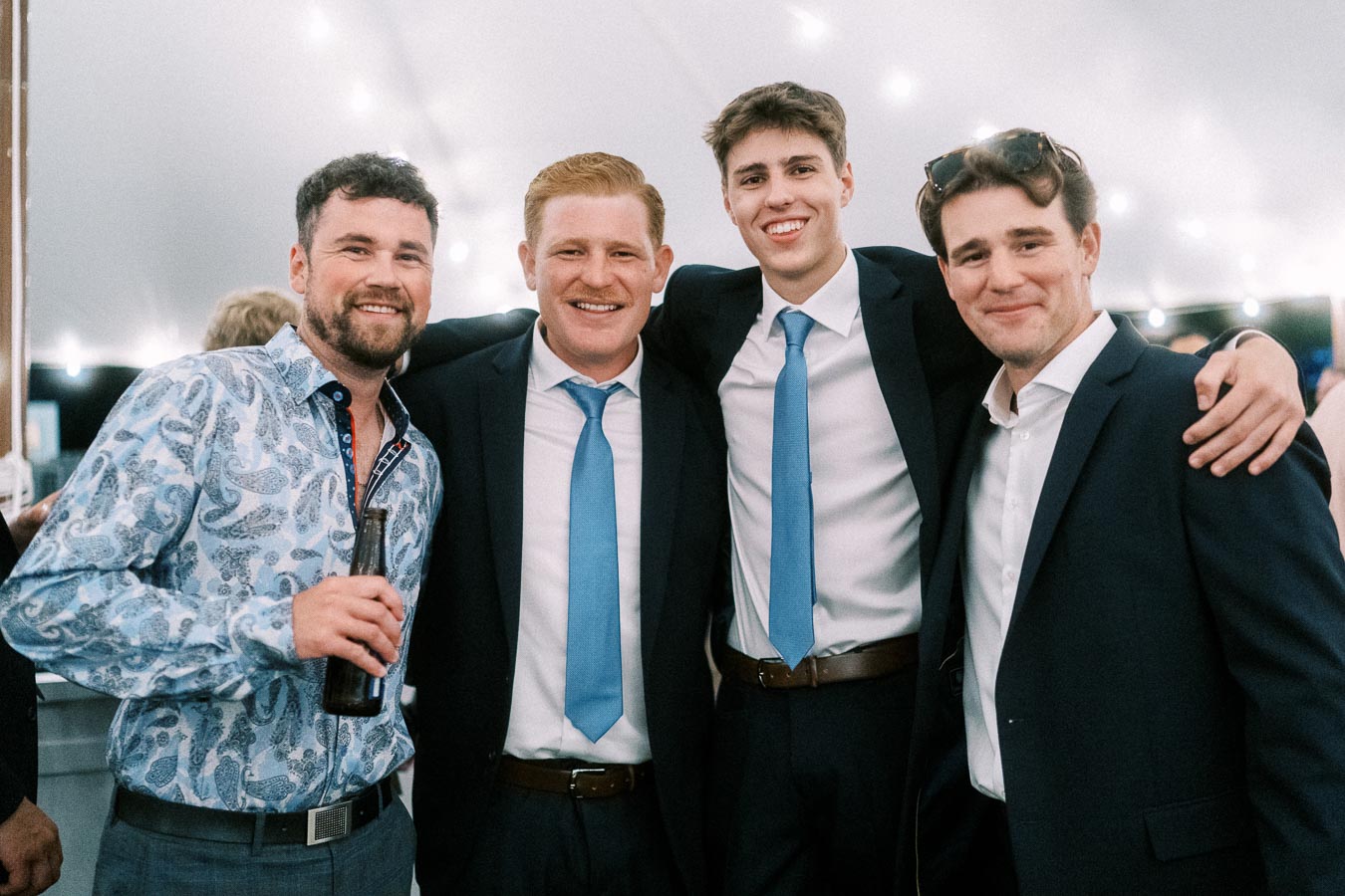 Four men smiling together under a wedding reception tent near Chatham-Kent, Ontario, with suits, blue ties, string lights, and a bottle in hand.