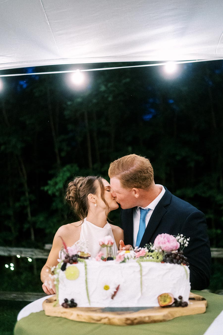 Bride and groom kissing behind a decorated wedding cake during an outdoor tented farm wedding reception near Chatham-Kent, Ontario, Canada.