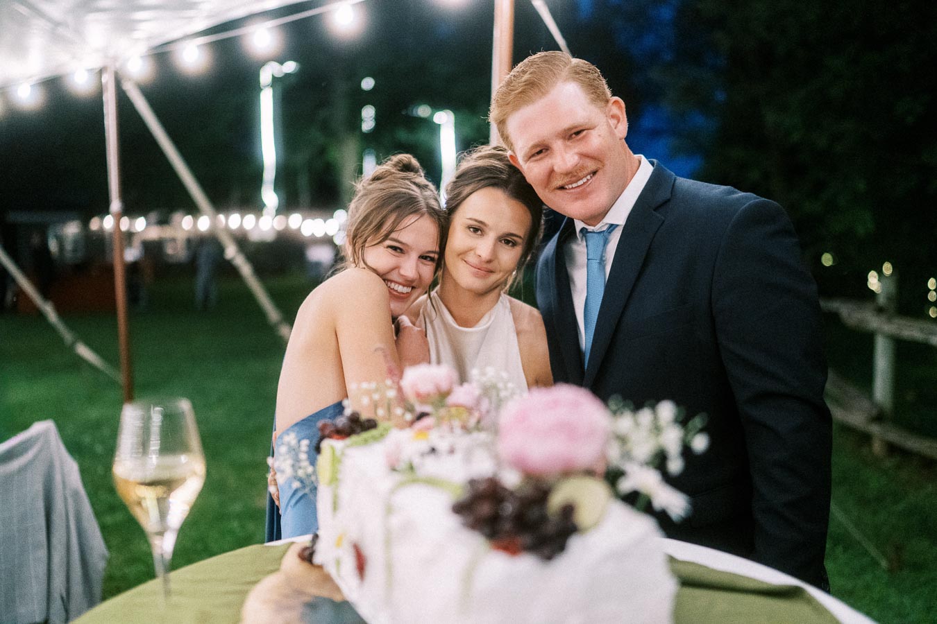 Three wedding guests smiling under a lit reception tent at an outdoor farm wedding near Chatham-Kent, Ontario, with flowers and wine on the table.