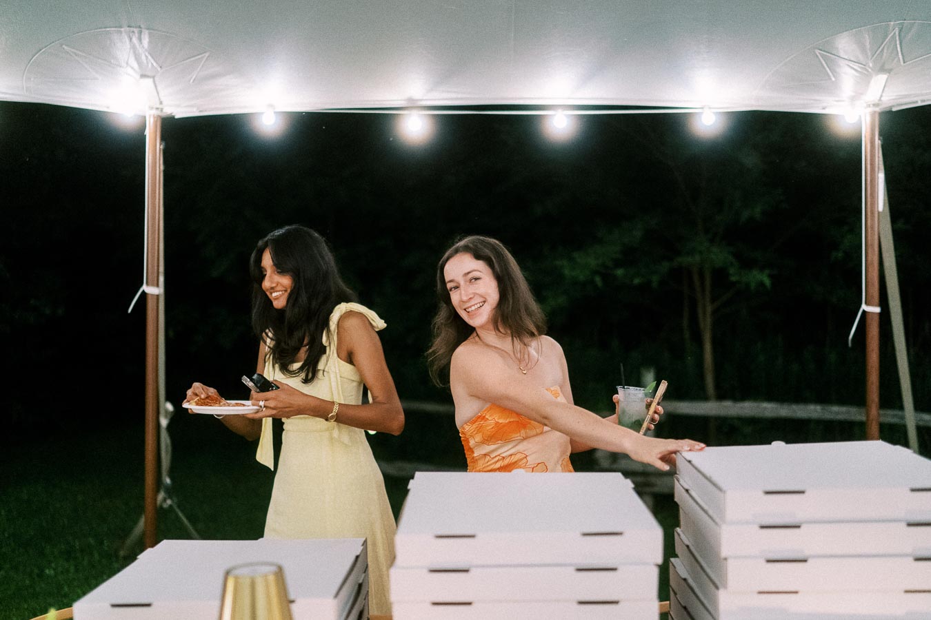 Two women at an outdoor tented farm wedding reception near Chatham-Kent, Ontario, with pizza boxes, drinks, and string lights at night.