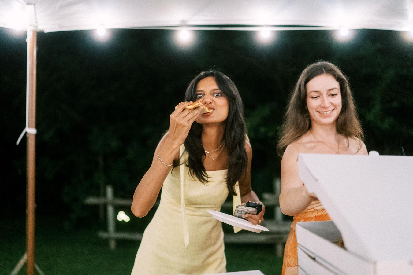 Two women enjoying pizza under a lighted outdoor wedding tent at night near Chatham-Kent, Ontario, Canada.